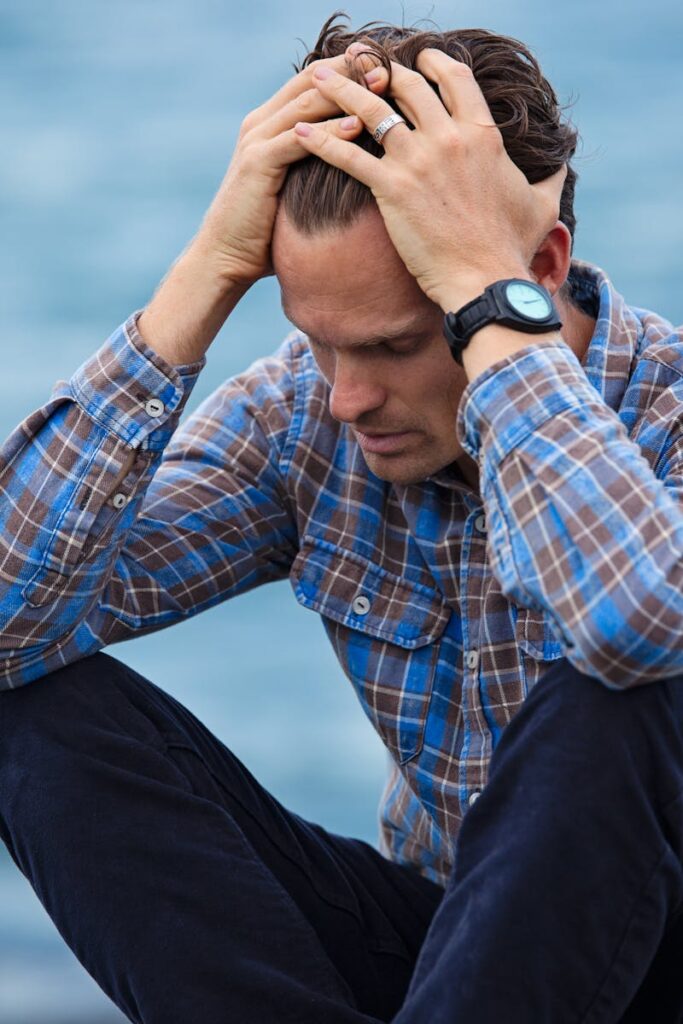 pexels photo 897817 897817 A man in a plaid shirt sits by the water looking distressed, symbolizing stress.
